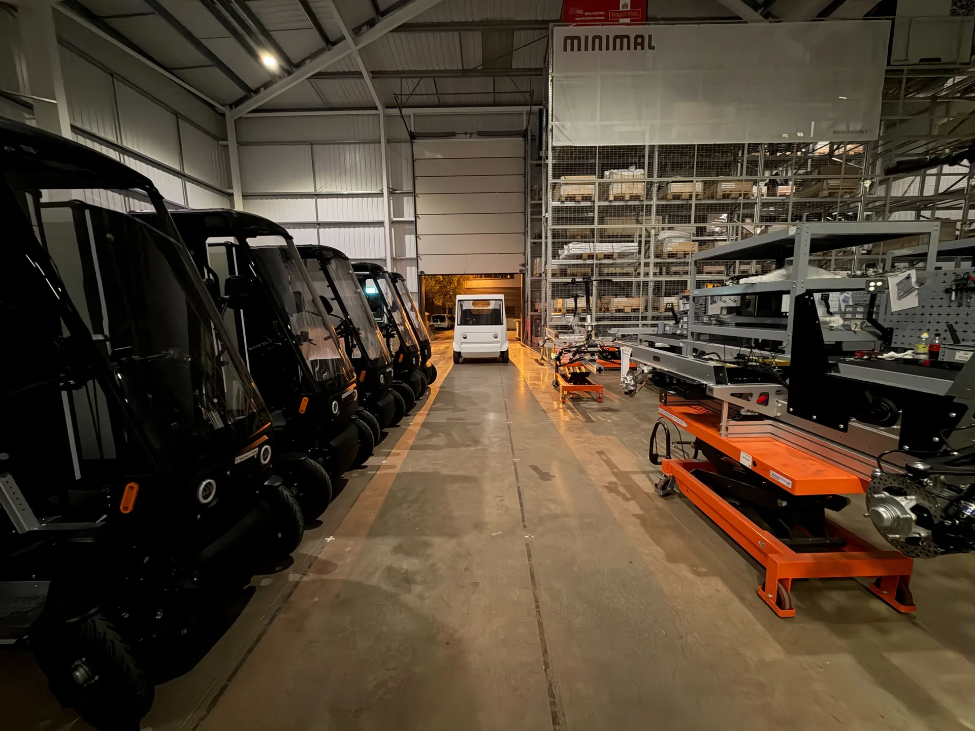 A row of Minimal vehicles parked nose to tail inside the factory, with assembly benches and orange trolleys along the far wall beneath a Minimal sign