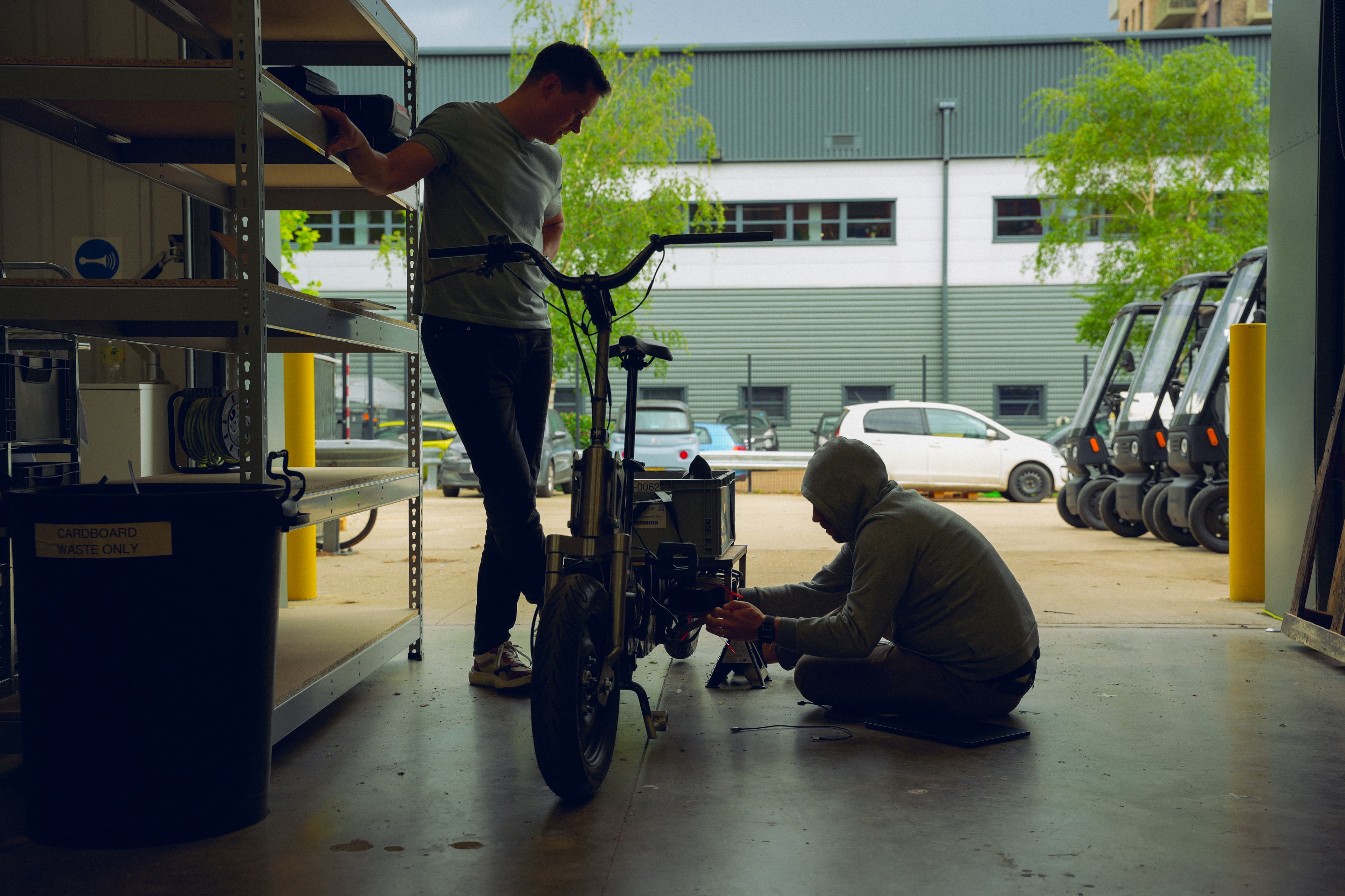 Two Minimal engineers servicing an electric vehicle in the workshop yard, with a row of vehicles parked behind